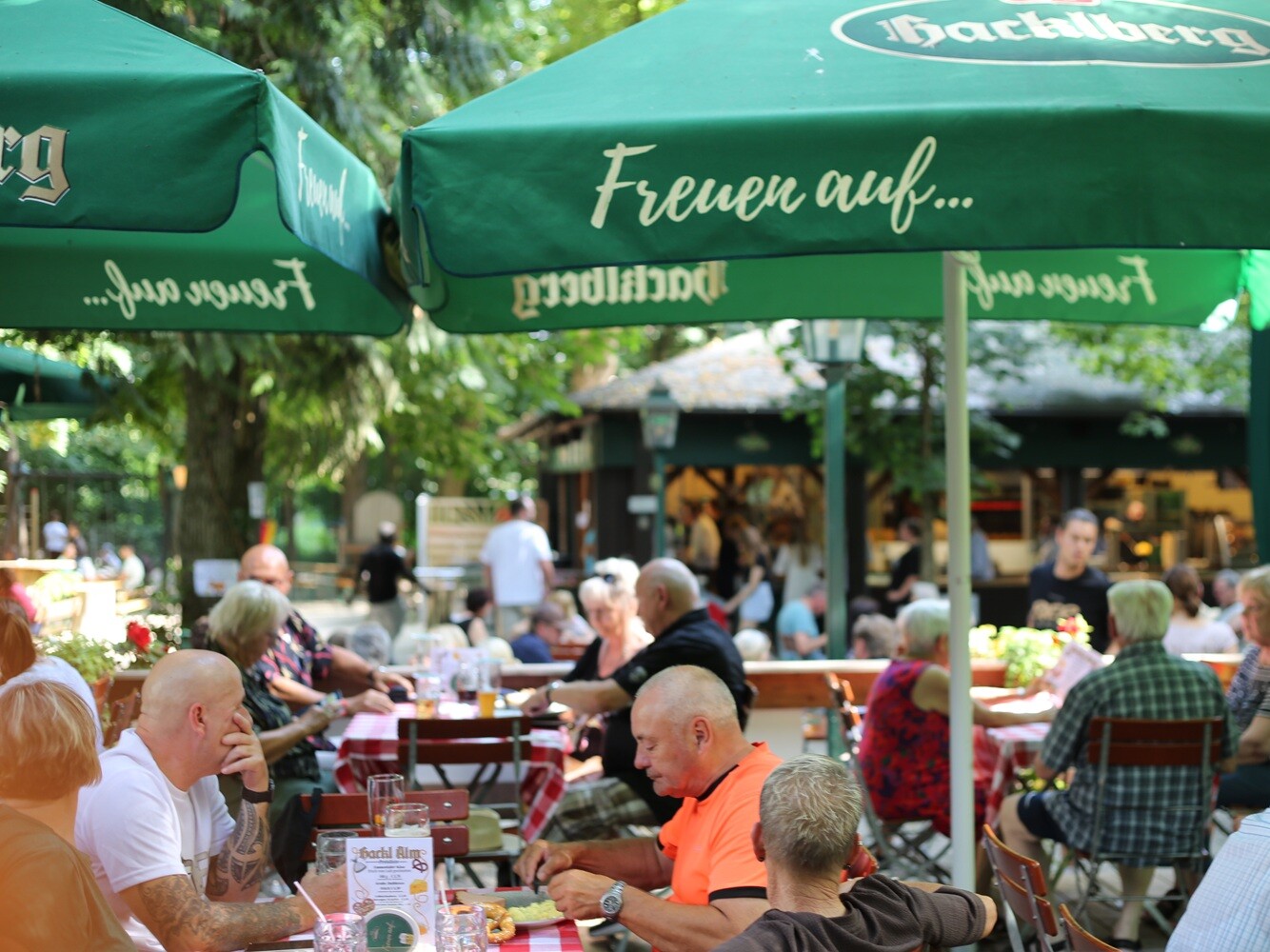 Gäste im Biergarten Eine Gruppe von Menschen sitzt an Tischen unter Sonnenschirmen.