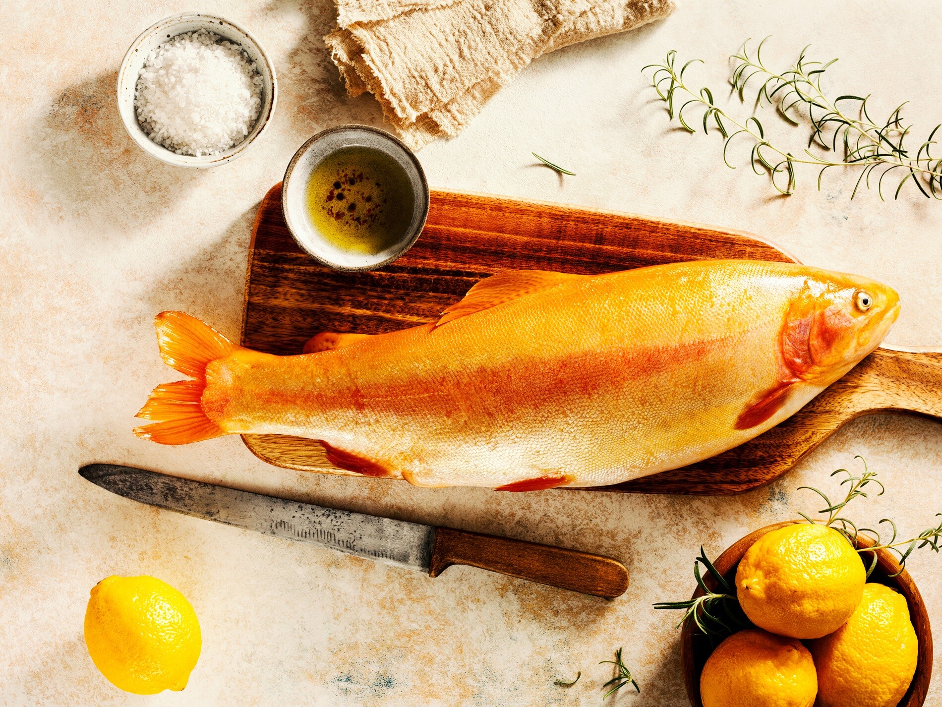 Frischer Fisch als Qualitätsmerkmal bei Bierbichler Rosenheim: Ein roher Fisch liegt auf einem Holzschneidebrett, daneben frische Zitronen und eine Schale mit Kräutern. Bierbichler bietet regionalen Süßwasserfisch sowie frische Salzwasserfische an. Ergänzend steht das vielfältige Fischsortiment auch als Tiefkühlware für Gastronomie, Hotellerie und Gemeinschaftsverpflegung zur Verfügung.